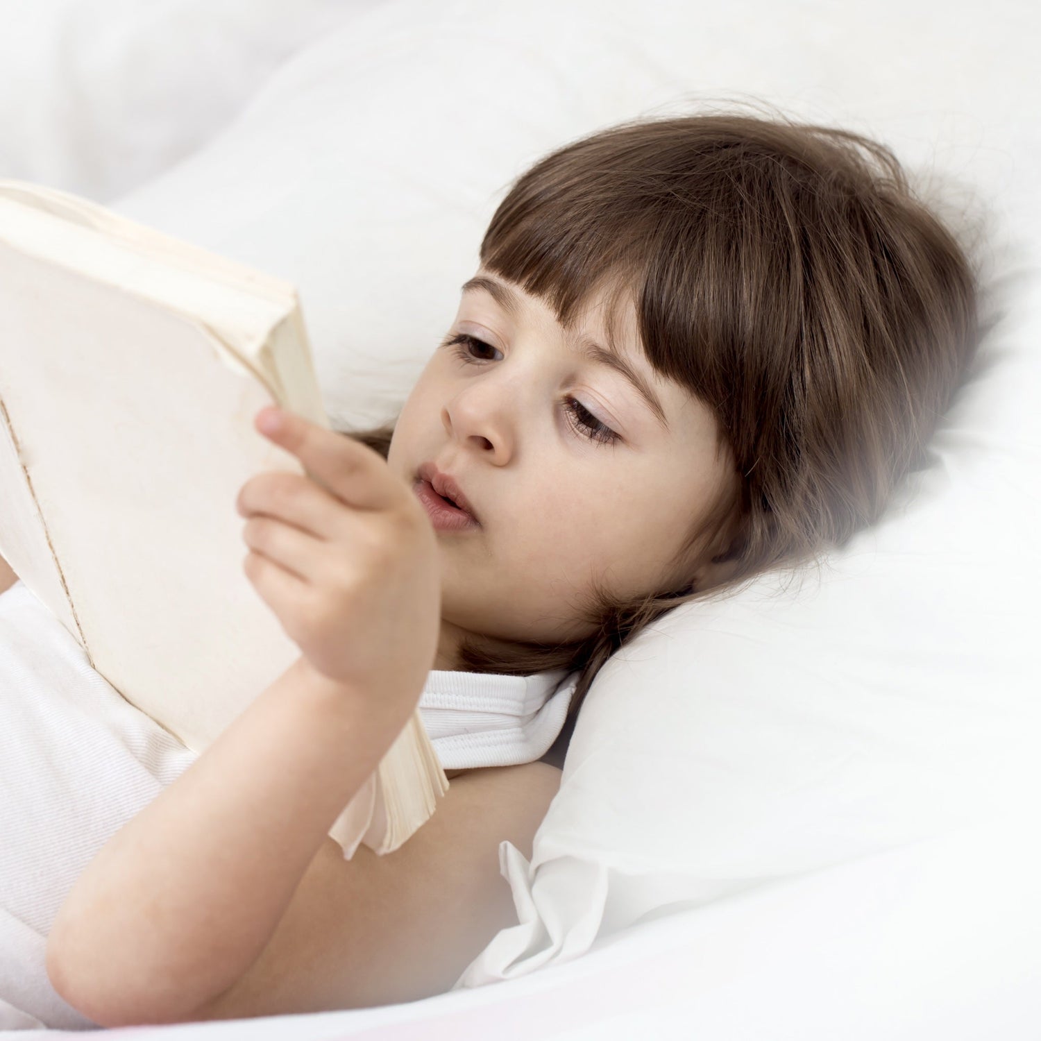 A young girl peacefully reading a book in bed, surrounded by the comfort of an Aurora Lilot Kids Balanced Pillow.