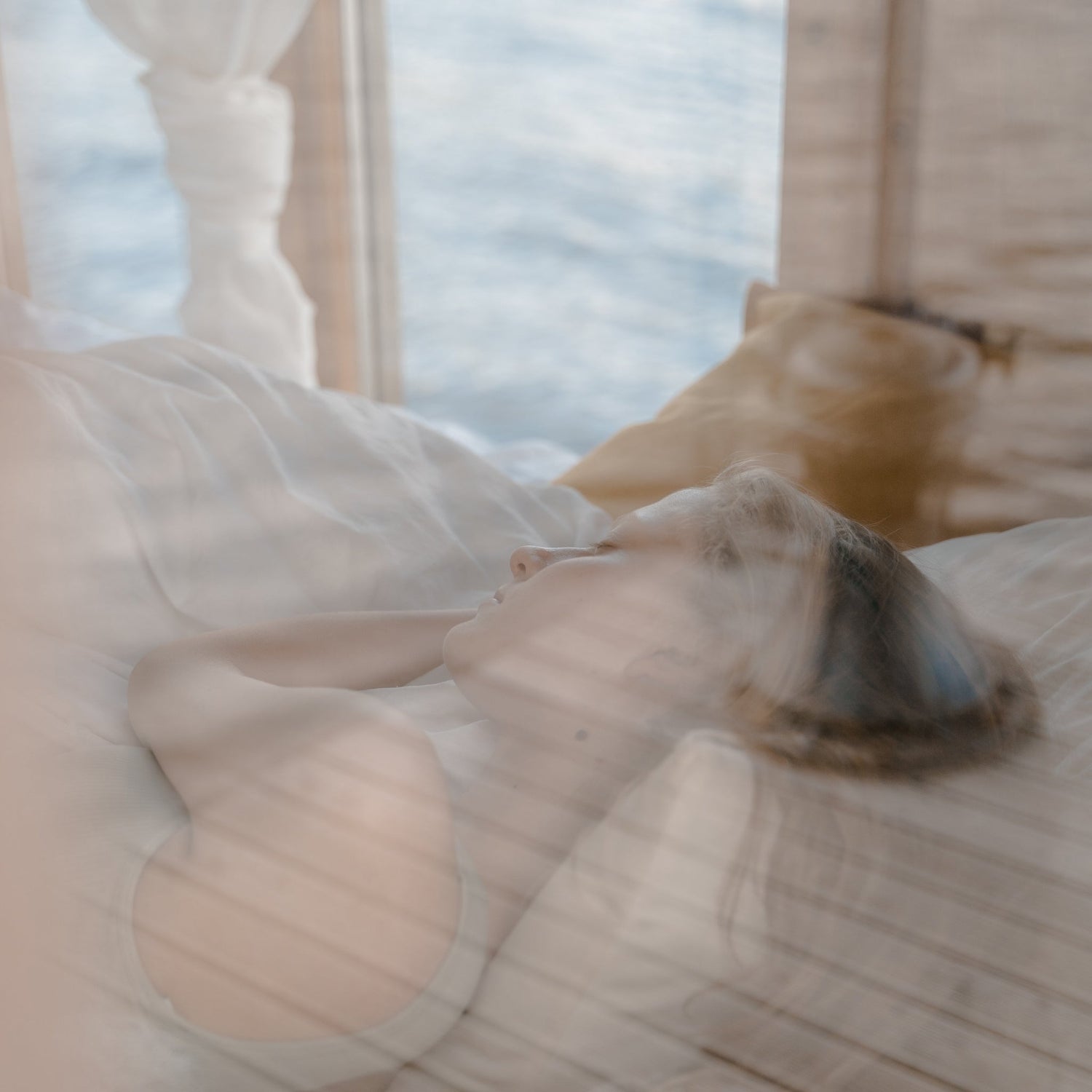 A beautiful woman sleeping sweetly, wrapped in the luxury Aurora Lilot Down Duvet and resting on a Lilot Down Pillow, with a serene view of the sea visible from her window. Atmosphere of tenderness and comfort.