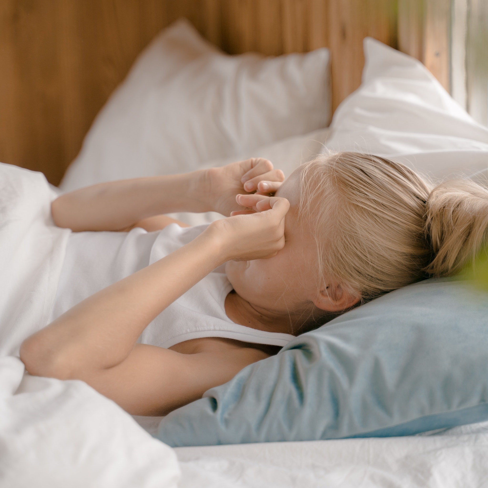 A young blonde girl lying in bed on a soft blue pillow, rubbing her eyes while surrounded by cozy white Aurora Lilot bedding, with a wooden wall in the background.